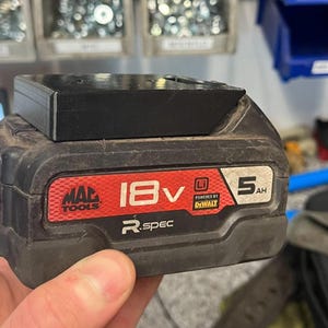 May include: A black and red 18V MAC TOOLS R.spec power tool battery with a black accessory attached. The battery is labeled "5AH" and "POWERED BY DEWALT". The background includes storage bins and other tools.