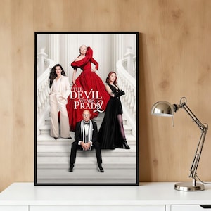 May include: A framed movie poster for "The Devil Wears Prada 2" featuring four people on a staircase. The poster has a black frame and the text "THE DEVIL WEARS PRADA 2" in the center. The people are wearing formal attire.