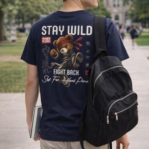 Op de afbeelding: Marineblauw T-shirt met een afbeelding van een vechtsport-teddybeer en de tekst "STAY WILD" en "FIGHT BACK". De beer draagt een hoofdband en een vechtsportuniform. Een zwarte rugzak is ook zichtbaar.