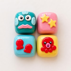 May include: Four colorful, square-shaped decorative objects. One is teal with a cartoon face, another is pink with yellow stars, a third is light blue with red shapes, and the last is yellow with a red octopus.