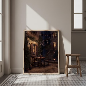 May include: A framed print of a European street scene at night. The image depicts a small table set for two, with chairs, in a cobblestone courtyard. String lights and a staircase are visible in the background.