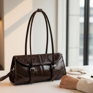 Puede incluir: Un bolso de mano de cuero marrón oscuro con asas largas y finas. El bolso tiene forma rectangular con cierre de solapa y dos hebillas plateadas. El cuero tiene un acabado brillante. El bolso está sobre una superficie blanca.