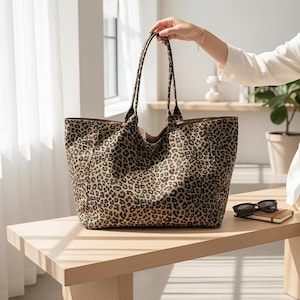 Op de afbeelding: Een grote tote bag met een luipaardprint. De tas wordt door een hand vastgehouden. De tas staat op een lichtgekleurde houten tafel. Zonnebril en een klein notitieboekje liggen ook op de tafel.