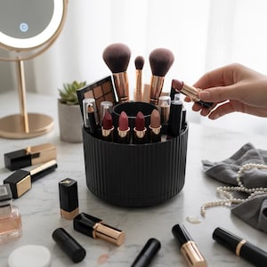 Puede incluir: Un organizador de maquillaje negro acanalado lleno de pintalabios, brochas y sombras de ojos. Una mano sostiene un pintalabios. Otros artículos de maquillaje están esparcidos sobre la superficie de mármol. Un espejo iluminado y una pequeña planta están en el fondo.