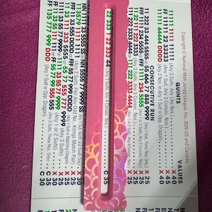 Puede incluir: Una tarjeta de la National Mah Jongg League 2026 para EE. UU. y Canadá con columnas de números y texto en tinta negra, verde y roja, parcialmente cubierta por una herramienta rectangular de plástico rosa con una ranura central y borde festoneado.