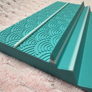May include: A rectangular, turquoise-colored object with a wave pattern on the top surface. A white, cylindrical rod is placed diagonally across the top. The object has a stepped design on one side, with white accents.