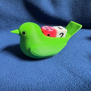 May include: A bright lime green 3D-printed bird figurine with a hollow back containing two dice. One die is translucent pink with black pips, and the other is a white cube with the black number 2 and the letter L clearly visible on its side.