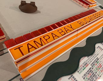 Puede incluir: Un soporte para fichas de juego de los Tampa Bay Buccaneers con un panel frontal de color naranja brillante con el nombre del equipo en letras negras, acompañado de una fila de fichas rojas translúcidas y una pequeña pieza de juego marrón.