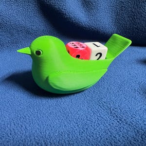 May include: A bright green 3D-printed bird figurine with a hollow back containing two dice. One die is translucent pink with black pips, and the other is a white cube with the black number 2 and the black letter L clearly visible on its side.