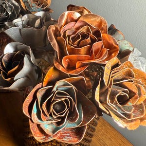 May include: A decorative arrangement of metal roses in shades of copper, bronze, and silver. The flowers are arranged in a textured, bronze-colored vase. The roses have intricate details and varying patinas, creating a unique and artistic display.