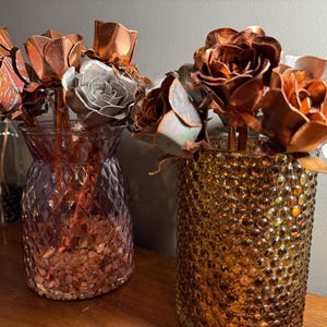 May include: Two decorative vases holding metal rose sculptures. One vase is a textured amber color, the other is a textured purple color. The metal roses are copper, silver, and bronze in color. The vases are on a wooden surface.