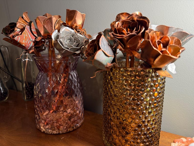 May include: Two decorative vases holding metal rose sculptures. One vase is a textured amber color, the other is a textured purple color. The metal roses are copper, silver, and bronze in color. The vases are on a wooden surface.
