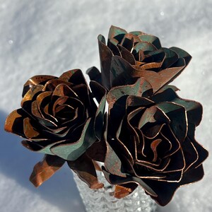 May include: A bouquet of three metal roses with a copper and verdigris finish. The handcrafted flowers are arranged in a textured, silver-colored vase, set against a snowy background. The roses have detailed petals and a rustic appearance.