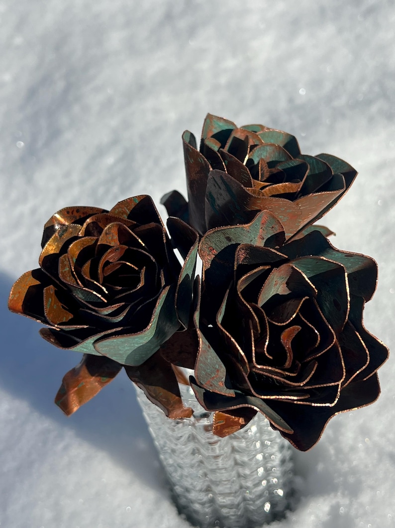 May include: A bouquet of three metal roses with a copper and verdigris finish. The handcrafted flowers are arranged in a textured, silver-colored vase, set against a snowy background. The roses have detailed petals and a rustic appearance.