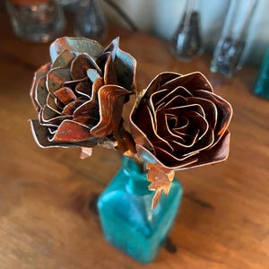 May include: A small bouquet of metal roses in a teal-colored square vase. The roses are made of copper, with a mix of brown and teal hues. The vase sits on a wooden surface, with other decorative glass bottles in the background.