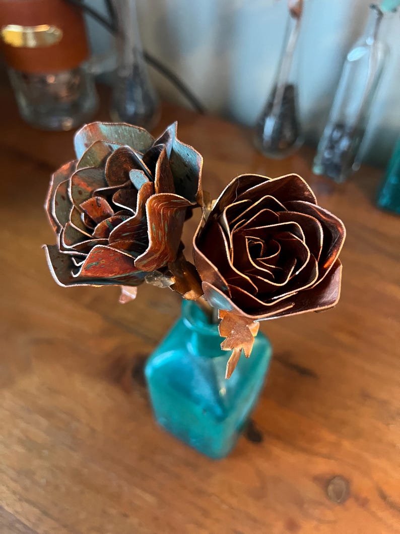 May include: A small bouquet of metal roses in a teal-colored square vase. The roses are made of copper, with a mix of brown and teal hues. The vase sits on a wooden surface, with other decorative glass bottles in the background.