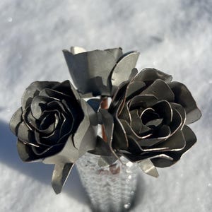 May include: A small bouquet of metal roses in a textured silver vase. The roses are crafted from dark gray metal, with detailed petals. The vase sits in a bed of snow, creating a contrast of textures and colors.