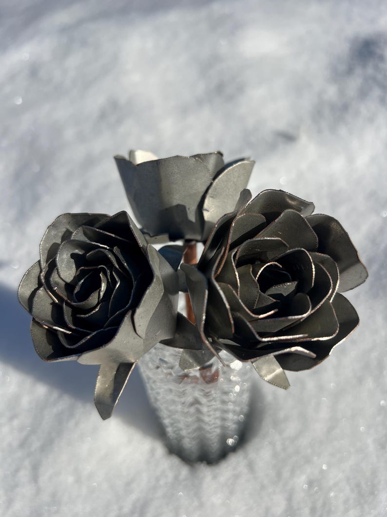 May include: A small bouquet of metal roses in a textured silver vase. The roses are crafted from dark gray metal, with detailed petals. The vase sits in a bed of snow, creating a contrast of textures and colors.