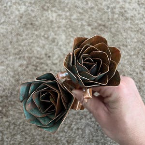 May include: Two handcrafted copper roses with a natural patina. The petals have a layered design, showcasing shades of brown and teal. The roses are connected by a copper stem, creating a unique floral sculpture.
