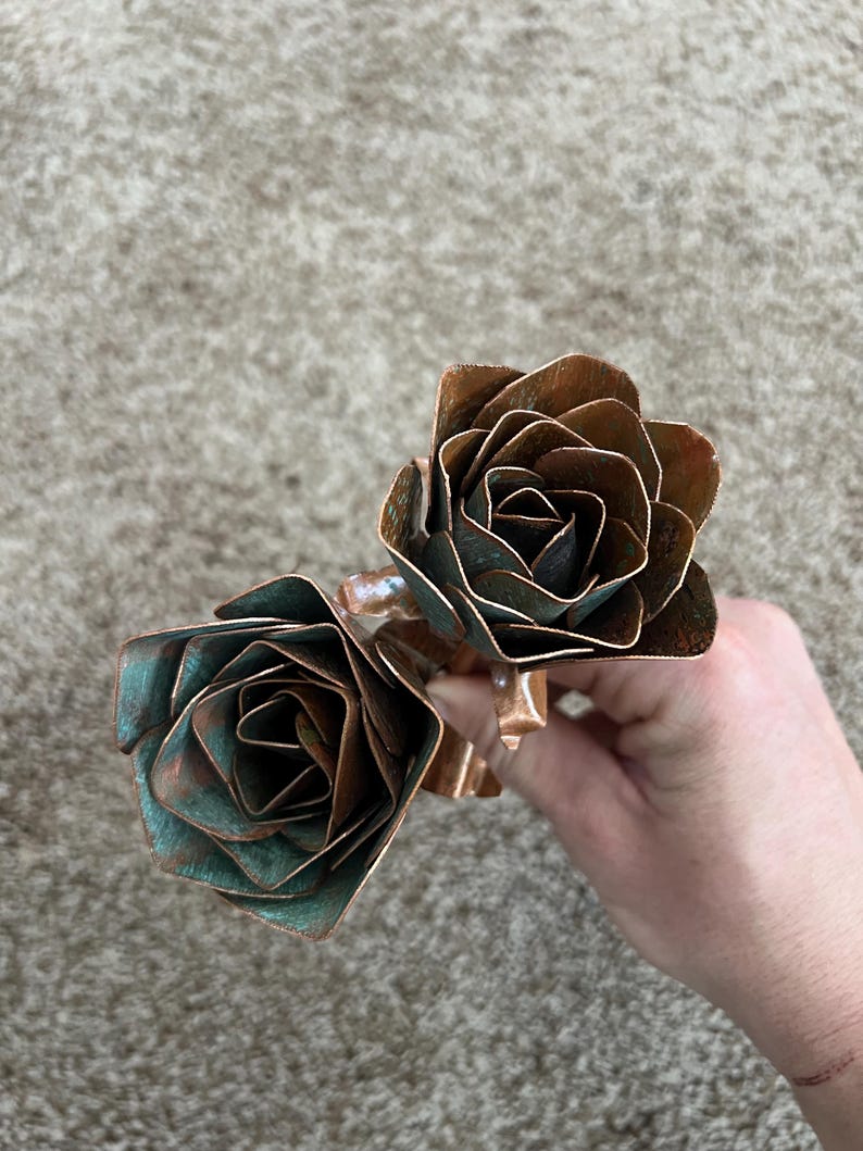 May include: Two handcrafted copper roses with a natural patina. The petals have a layered design, showcasing shades of brown and teal. The roses are connected by a copper stem, creating a unique floral sculpture.