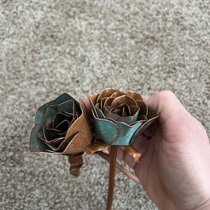 May include: Two metal rose sculptures with copper and teal patina. The roses are held by a copper stem. The petals are detailed and layered, creating a realistic appearance. The roses are a unique decorative item.