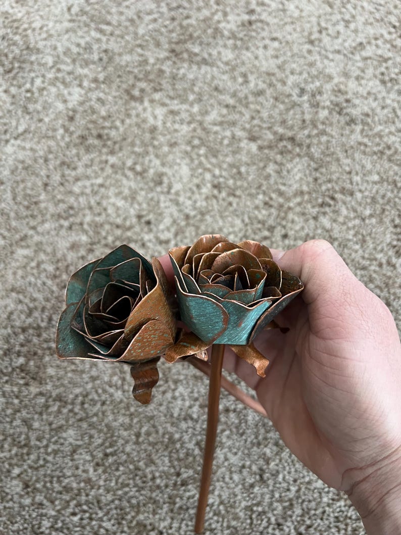 May include: Two metal rose sculptures with copper and teal patina. The roses are held by a copper stem. The petals are detailed and layered, creating a realistic appearance. The roses are a unique decorative item.
