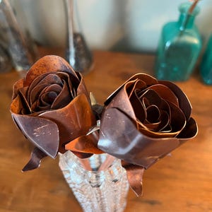 May include: Two handcrafted metal roses in a clear, textured glass vase. The roses are a rich, dark brown with detailed petal construction. The vase sits on a wooden surface, with blurred glass bottles in the background, suggesting a decorative floral arrangement.