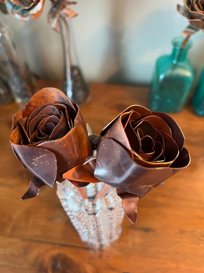 May include: Two handcrafted metal roses in a clear, textured glass vase. The roses are a rich, dark brown with detailed petal construction. The vase sits on a wooden surface, with blurred glass bottles in the background, suggesting a decorative floral arrangement.
