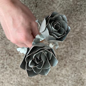 May include: Two silver metal roses with detailed petal construction. The roses are held in a hand, showcasing their metallic sheen and intricate design. The flowers are a unique decorative item.