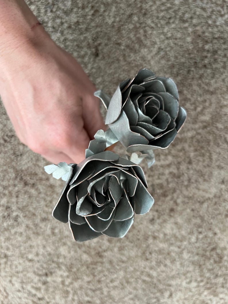 May include: Two silver metal roses with detailed petal construction. The roses are held in a hand, showcasing their metallic sheen and intricate design. The flowers are a unique decorative item.