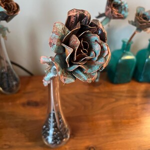 May include: A handcrafted copper rose with a verdigris finish, displayed in a clear glass vase. The rose petals have a brown and teal patina. Several other copper roses are visible in the background, some in teal glass bottles.