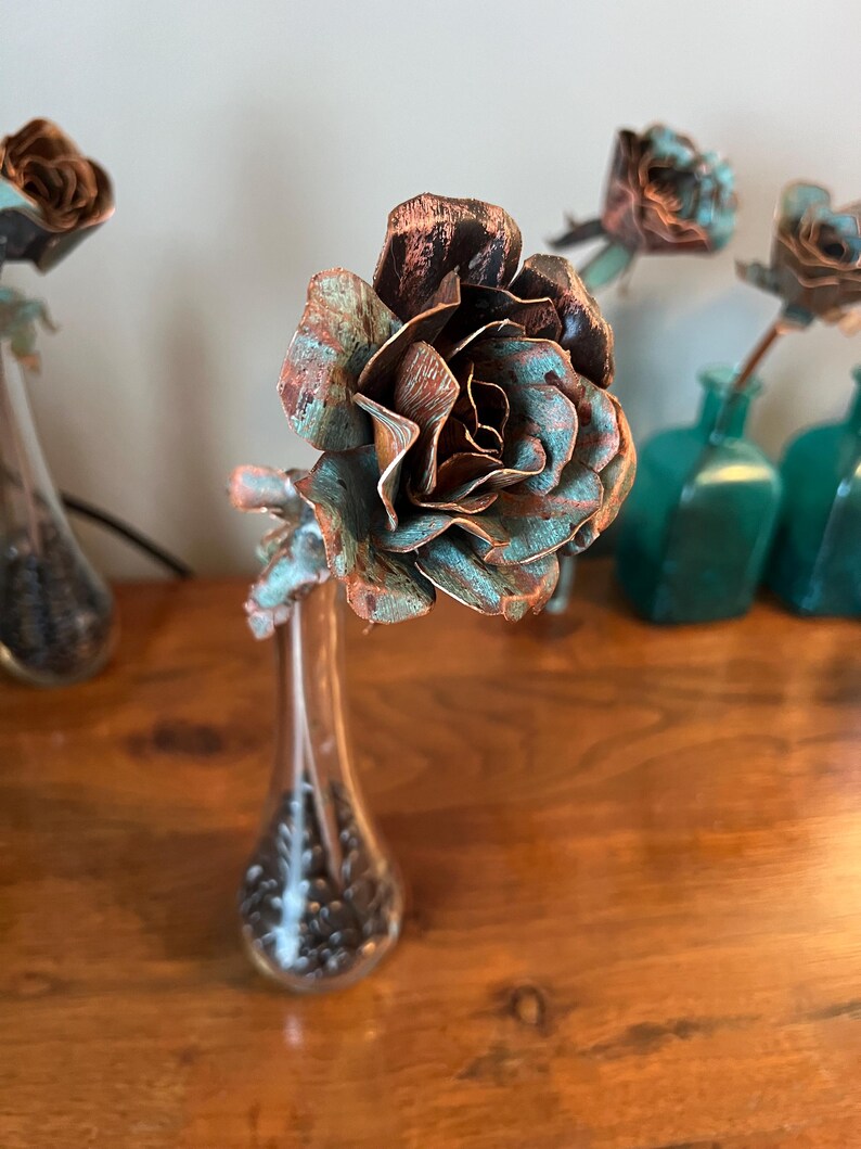 May include: A handcrafted copper rose with a verdigris finish, displayed in a clear glass vase. The rose petals have a brown and teal patina. Several other copper roses are visible in the background, some in teal glass bottles.