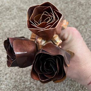 May include: Three handcrafted copper roses with detailed petal construction. The roses display varying shades of brown and copper, with a metallic sheen. The stems are also copper, creating a cohesive floral arrangement.