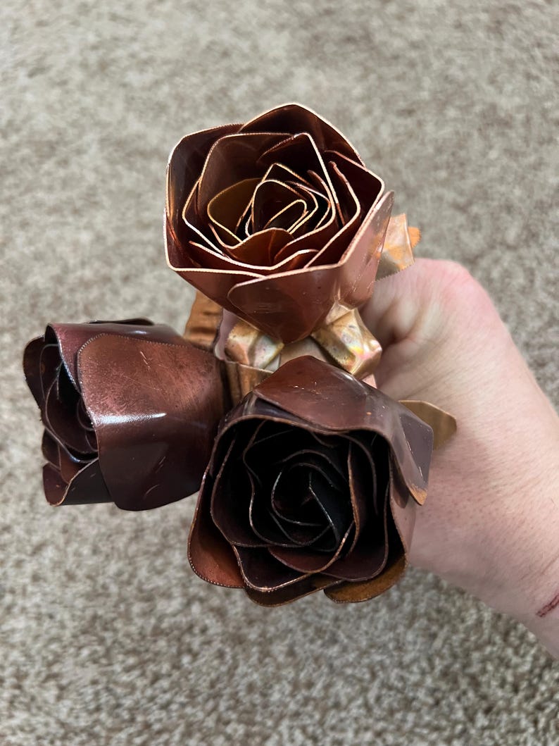 May include: Three handcrafted copper roses with detailed petal construction. The roses display varying shades of brown and copper, with a metallic sheen. The stems are also copper, creating a cohesive floral arrangement.