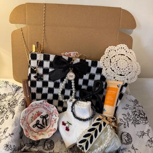 Peut inclure: Coffret cadeau garni de divers articles : pochette à carreaux noirs et blancs, petit plat fleuri, tube de gloss, crème pour les mains, chaussettes brodées de cerises et plusieurs bijoux. Un napperon et une étiquette "Merci" sont également présents.