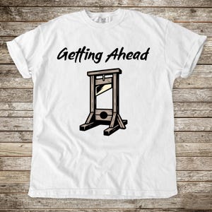 May include: White t-shirt with the phrase "Getting Ahead" above a cartoon illustration of a guillotine. The guillotine is depicted in brown and black, with a blade and a wooden frame. The shirt has short sleeves.