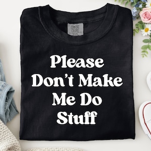 May include: Black t-shirt with the phrase "Please Don't Make Me Do Stuff" in white, bold, sans-serif font. The shirt has a crew neck and a tag that reads "Comfort Colors". The shirt is folded.