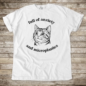 May include: White t-shirt with a black graphic of a cat's face and the text "full of anxiety and microplastics" in a circular layout. The cat has large eyes and striped fur. The shirt has short sleeves and a crew neck.
