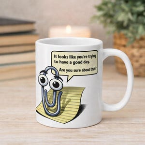 May include: White ceramic mug featuring a cartoon paperclip character with googly eyes and a speech bubble. The speech bubble reads, "It looks like you're trying to have a good day. Are you sure about that?" The mug has a curved handle.