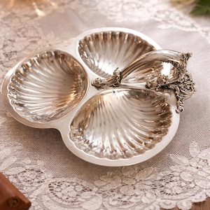 May include: Silver-toned serving dish with three shell-shaped compartments and a decorative handle. The dish has a scalloped design and sits on a lace tablecloth. The dish is likely used for serving appetizers or small portions.