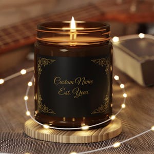 May include: A lit candle in a brown glass jar with a black label. The label has gold floral corner designs and gold text that reads "Custom Name Est. Year". The candle sits on a round wooden coaster, surrounded by string lights.