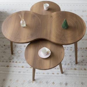 Puede incluir: Un conjunto de tres mesas de centro de madera con un diseño único de trébol. Las mesas son de color marrón cálido y tienen un acabado liso. Un pequeño árbol de Navidad decorativo, una taza y un difusor están colocados sobre las mesas.