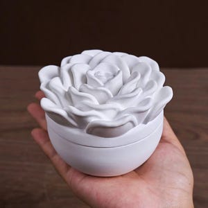 以下が含まれることがあります： 白いバラの形をした小物入れ。 蓋は、詳細な花びらを持つ、完全に咲いたバラに似せて彫刻されています。 ベースはシンプルで丸みを帯びた容器です。 箱は手で持たれています。