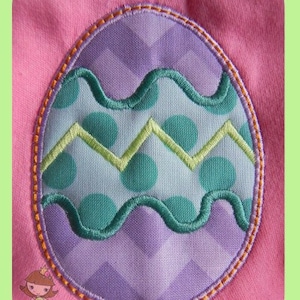 以下が含まれることがあります： ピンク色の生地に刺繍されたイースターエッグのデザイン。卵は紫色のシェブロン模様、ターコイズのドット、波状の緑色のジグザグ模様が特徴です。卵はオレンジ色のステッチで縁取られています。