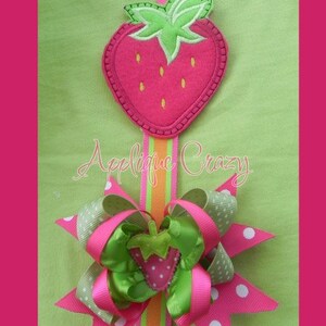 May include: A green, pink, and white bow with a strawberry applique in the center. The bow is attached to a green, orange, and white ribbon. The ribbon is attached to a strawberry applique with a green stem and leaves. The text "Applique Crazy" is visible in the image.