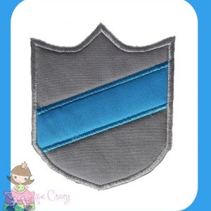 Può includere: Uno scudo in tessuto grigio con una striscia diagonale blu. Lo scudo ha un bordo cucito.