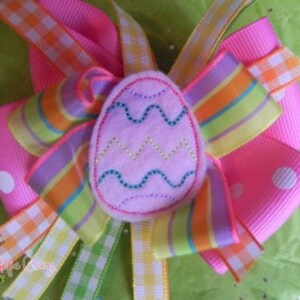 以下が含まれることがあります： 明るいピンク色のヘアリボン。中央にはフェルトのイースターエッグが飾られています。リボンには、水玉模様、ストライプ、ギンガムチェックなど、さまざまなリボンが使用されています。卵は薄いピンク色で、ジグザグ模様です。