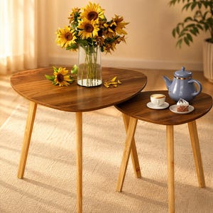 May include: Two wooden nesting tables with a triangular top and light-colored legs. A vase of sunflowers sits on the larger table. A blue teapot, teacup, and cake are on the smaller table. The tables are on a light-colored rug.
