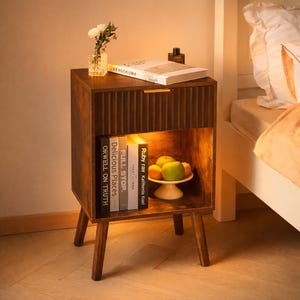 May include: A wooden nightstand with a ribbed drawer front and a lower shelf. The nightstand holds books, a small vase with flowers, and a bowl of fruit. The books include titles like "ORWELL ON TRUTH" and "FULL STOP."