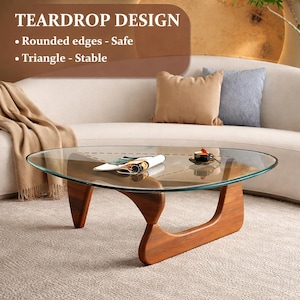 May include: A modern coffee table with a clear glass top and a unique wooden base. The table has a teardrop design with rounded edges and a stable triangle base. A magazine and a cup of coffee are on the table.